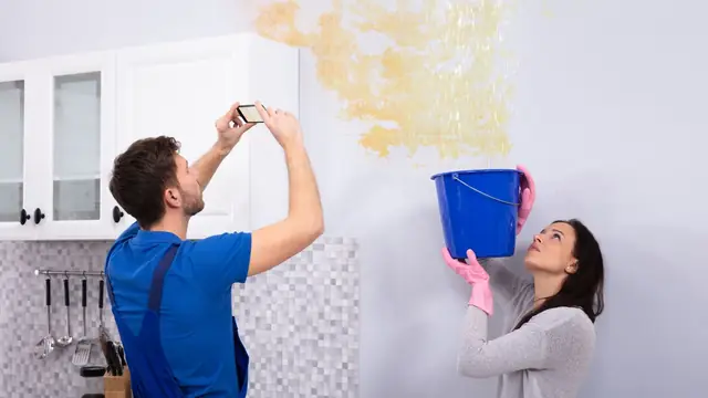 Homeowners photographing a leaking ceiling stain while catching drips in a bucket