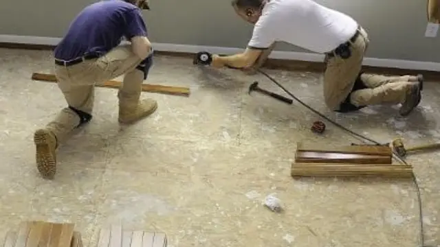 Two workers kneeling on a bare subfloor while cutting out damaged flooring